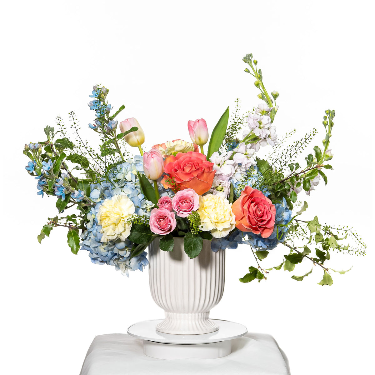 Radiance arrangement of blue delphinium, peach roses and white stock in a white footed urn
