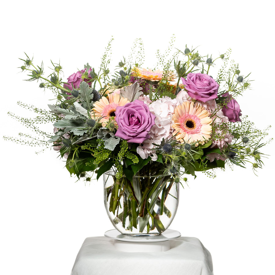 Exquisita arrangement of lilac roses, peach gerberas and dusty miller in a glass vase