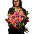 Amnesia bouquet of coral roses and magenta blooms with dark foliage, held by a florist