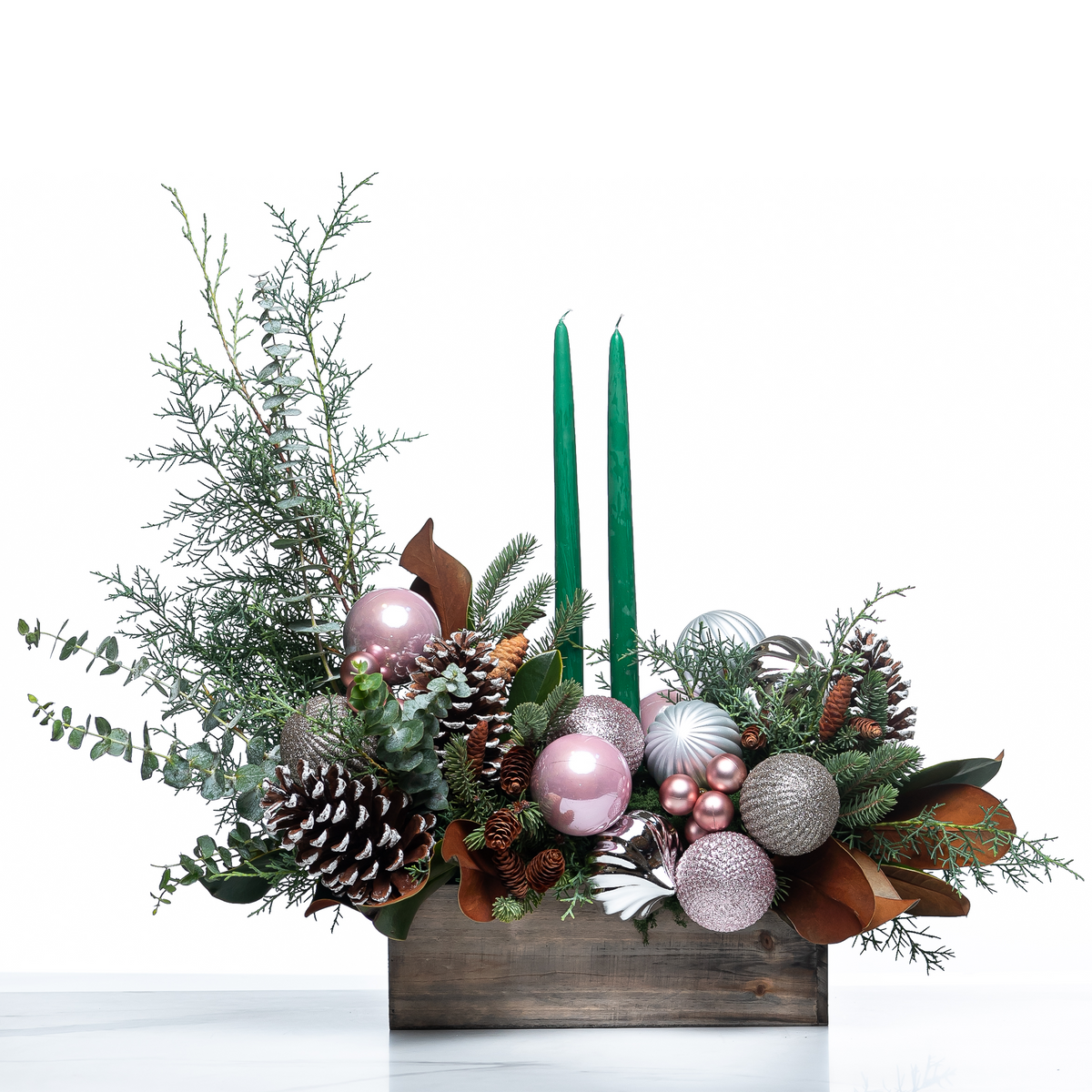 Blush and Balsam Glow arrangement with green taper candles, pink baubles and pine cones in a wooden box