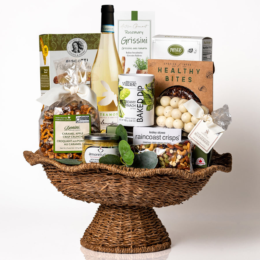 Heirloom Harvest: a wicker pedestal bowl with a wavy rim holding a bottle of Fonteamonte Trebbiano, a baked spinach dip, Raincoast Crisps, grissini, yogurt covered blueberries and biscotti