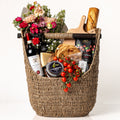 Marketta Marché: a tall woven seagrass basket with a wooden bar handle holding a fresh bouquet, a bottle of Chianti, a baguette, cherry tomatoes on the vine, My Mother's olives and olive oil, Parmigiano and a wooden board