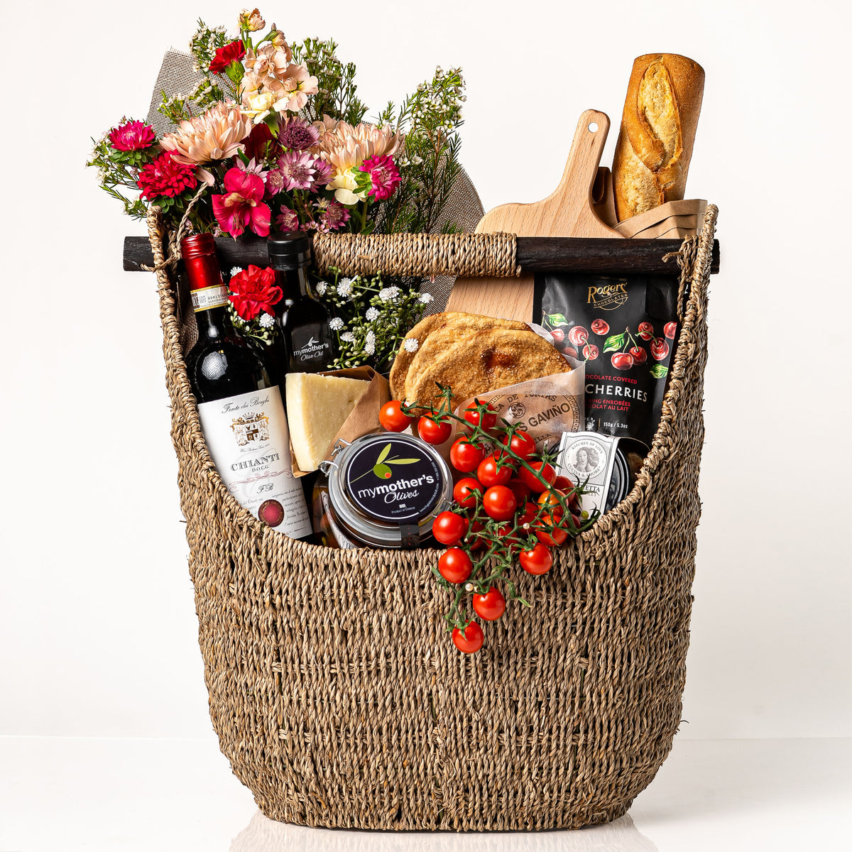 Marketta Marché: a tall woven seagrass basket with a wooden bar handle holding a fresh bouquet, a bottle of Chianti, a baguette, cherry tomatoes on the vine, My Mother's olives and olive oil, Parmigiano and a wooden board