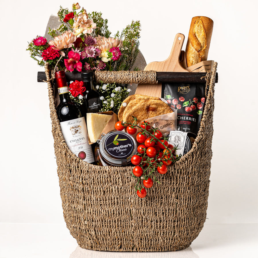 Marketta Marché: a tall woven seagrass basket with a wooden bar handle holding a fresh bouquet, a bottle of Chianti, a baguette, cherry tomatoes on the vine, My Mother's olives and olive oil, Parmigiano and a wooden board