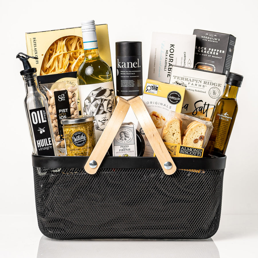Pantry Provisions: a black metal market basket with wooden handles holding a bottle of Pinot Grigio, egg tagliatelle, My Mother's olive oil, an oil and vinegar bottle, Kanel peppercorns, pesto, tapenade, crackers, pistachios and almond biscotti