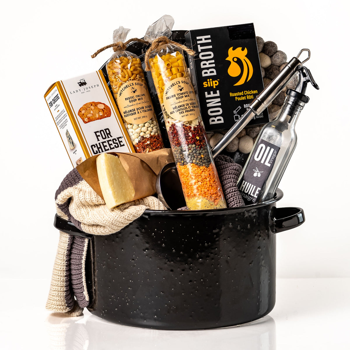 Pot of Plenty: a black enamel stock pot holding layered soup mixes, roasted chicken bone broth, a stainless steel ladle, an oil and vinegar bottle, knit tea towels, a felt ball trivet, cheese crisps and a Parmigiano wedge