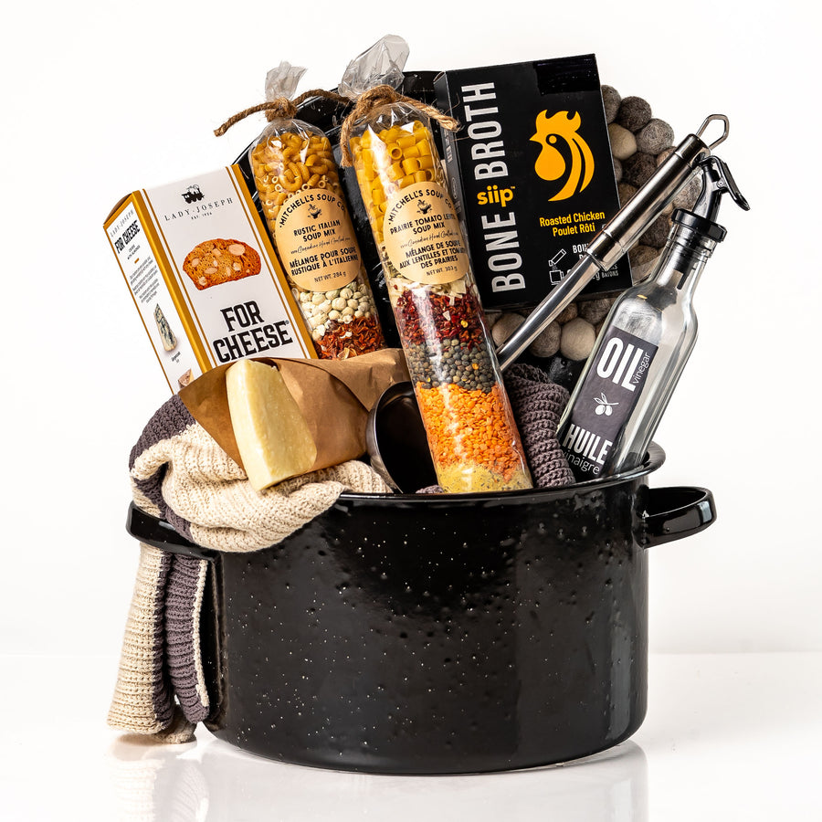 Pot of Plenty: a black enamel stock pot holding layered soup mixes, roasted chicken bone broth, a stainless steel ladle, an oil and vinegar bottle, knit tea towels, a felt ball trivet, cheese crisps and a Parmigiano wedge