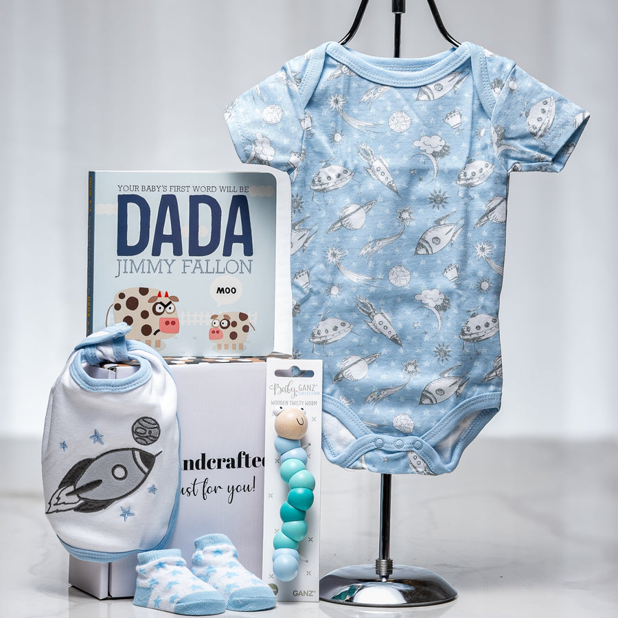 Soar baby boy gift with a blue patterned onesie on a hanger, the book Dada by Jimmy Fallon and booties