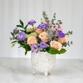 Sprouting arrangement of purple lisianthus, peach roses and eucalyptus in a speckled footed pot