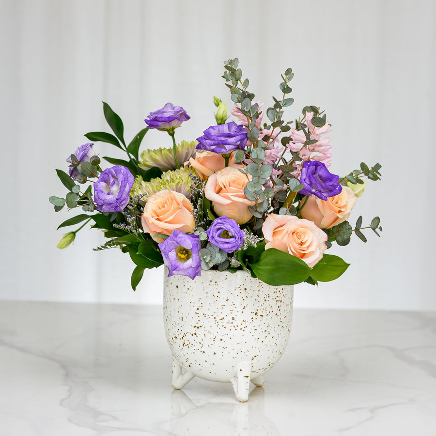 Sprouting arrangement of purple lisianthus, peach roses and eucalyptus in a speckled footed pot