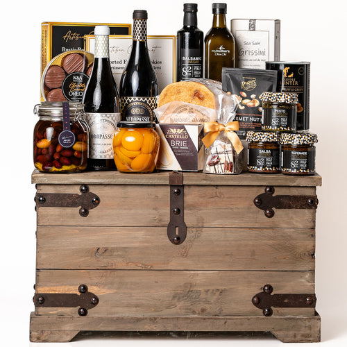 Treasure Trove Large: a rustic wooden trunk with black metal detailing overflowing with Biscardo wines, My Mother's olive oil and olives, balsamic vinegar, peach halves, chocolate covered Oreos, Castello brie and Co. 82 jars
