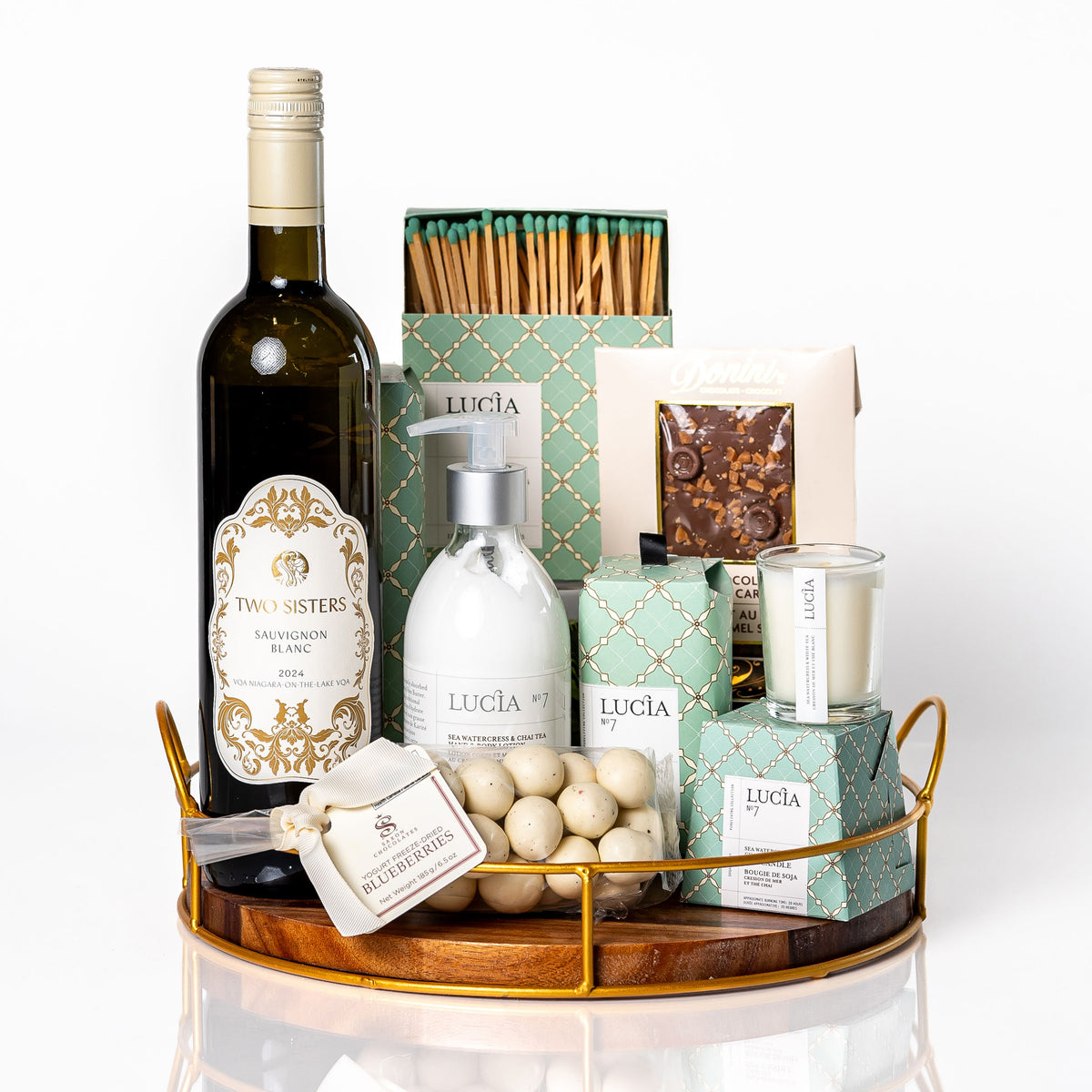Unwine'd: a round wood tray with gold edging holding a bottle of Two Sisters Sauvignon Blanc, Lucia No. 7 hand and body lotion, soy candle, bar soap and matches, yogurt covered blueberries and a Donini chocolate bar