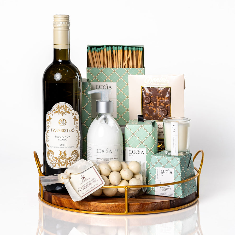 Unwine'd: a round wood tray with gold edging holding a bottle of Two Sisters Sauvignon Blanc, Lucia No. 7 hand and body lotion, soy candle, bar soap and matches, yogurt covered blueberries and a Donini chocolate bar