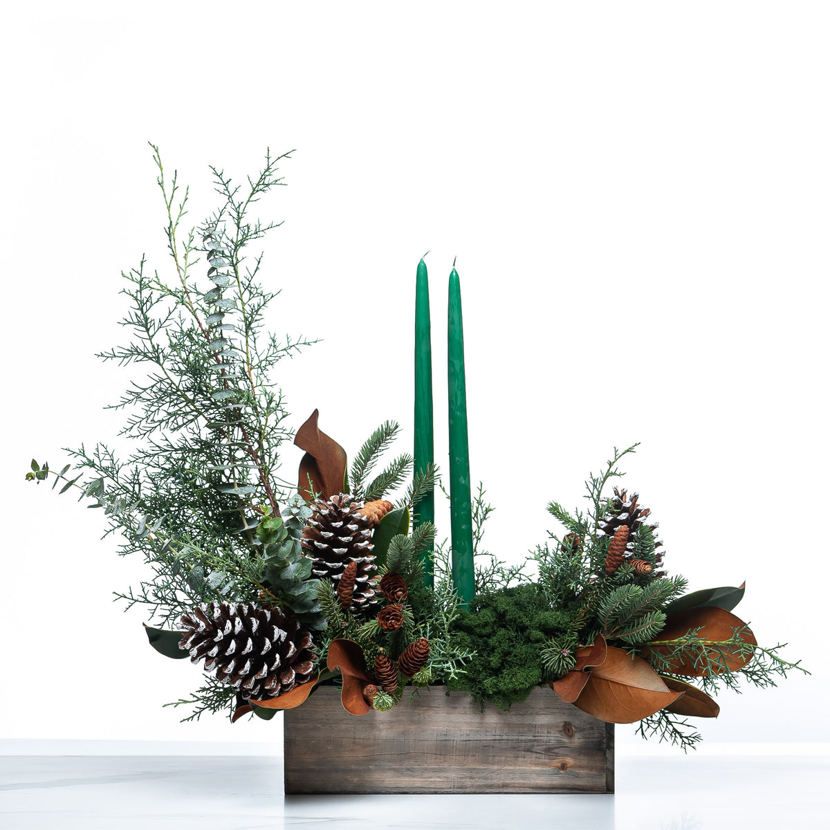 Woodland Grace and Glow arrangement with green taper candles, pine cones and magnolia in a wooden box