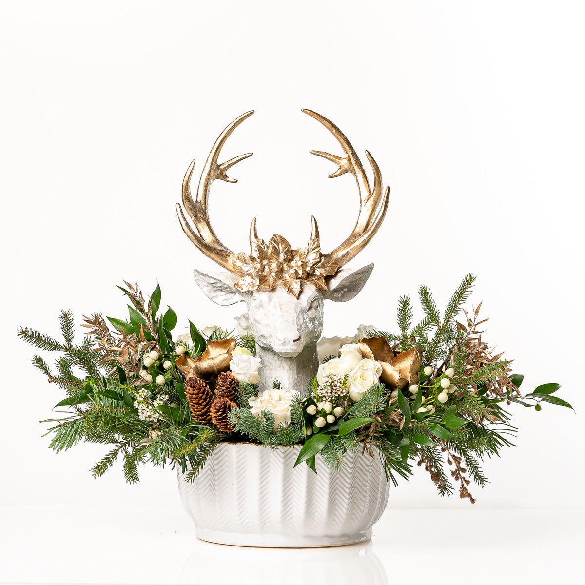 Woodland Wonder: a holiday floral centrepiece in a white fluted planter with a white deer head and gold antlers, surrounded by ivory roses, winter evergreens, white berries and pinecones
