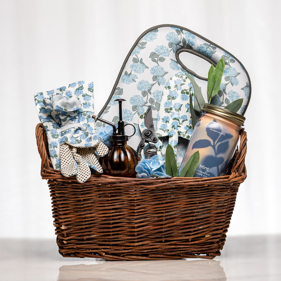 Hydrangea Haven gardening gift basket with blue hydrangea-patterned tool set, kneeling pad, gloves, glass plant mister and a herb growing kit in a lined wicker basket