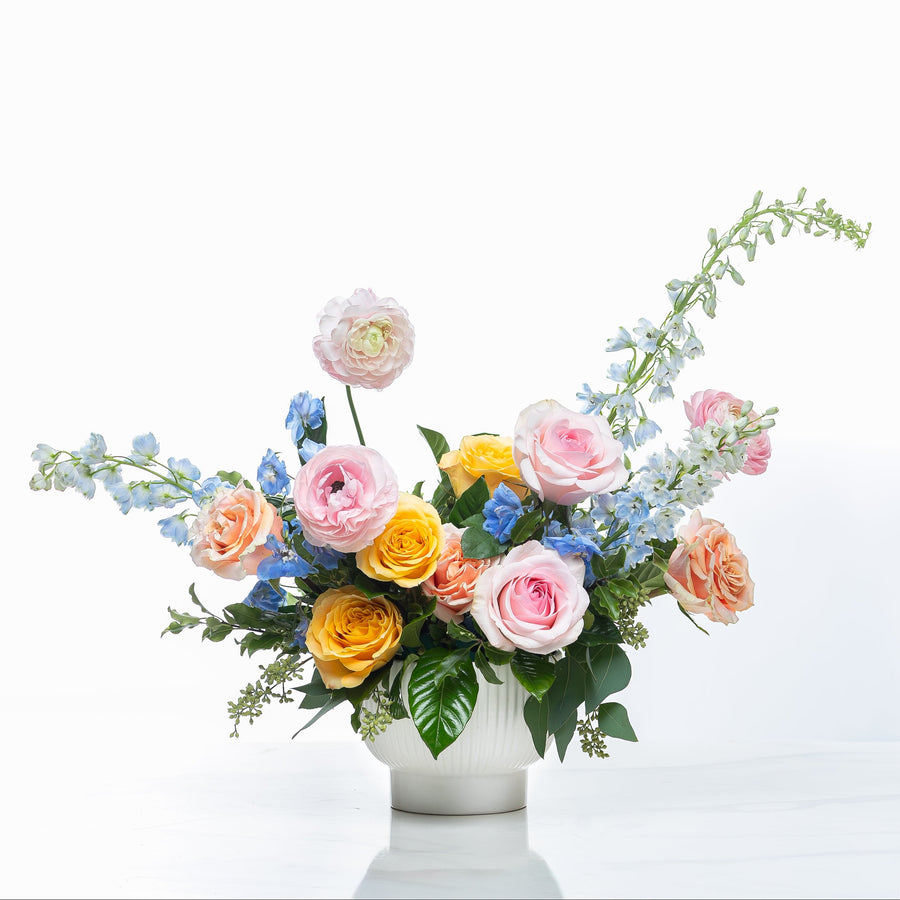 Whispering Petals arrangement of blue delphinium, peach roses and ranunculus in a white pot
