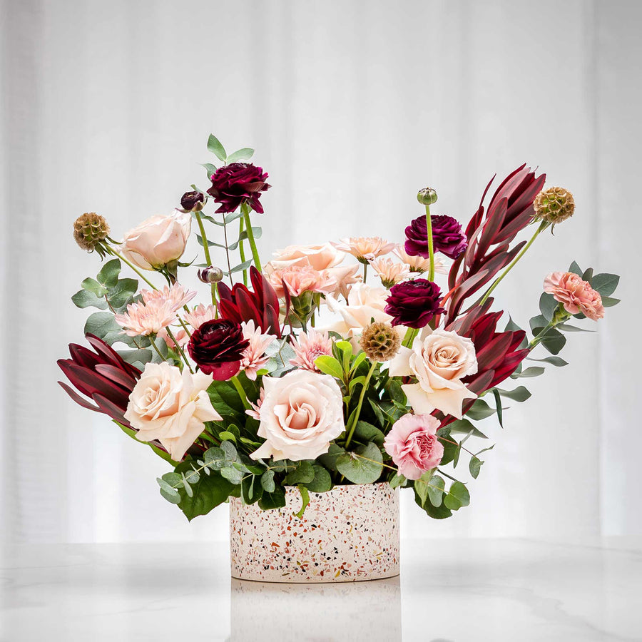 Bohemian Vision floral arrangement of blush and burgundy roses with protea in a speckled pink vase
