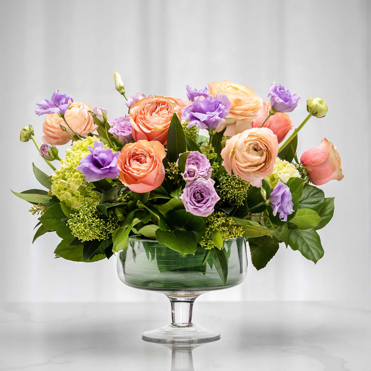 Macaroon floral arrangement of peach and lavender roses with green hydrangea in a footed glass bowl